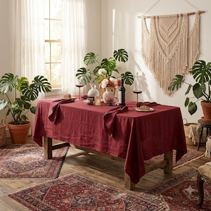 May include: A rectangular wooden dining table covered with a deep red linen tablecloth. The table is set with two wine glasses, a dark wine bottle, a white ceramic vase with flowers, and two small plates. The setting is surrounded by potted plants.