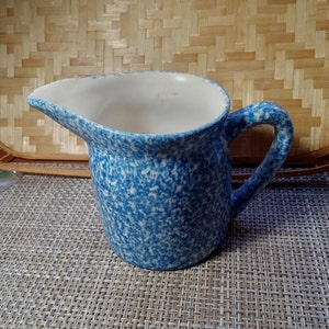 May include: A blue and white speckled ceramic pitcher with a handle. The pitcher is on a woven mat.