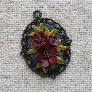 May include: An oval pendant with a dark bronze filigree frame. The pendant features a teal background with a cluster of burgundy flowers and yellow-green leaves. The pendant has a loop at the top for hanging.