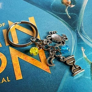 May include: A silver-toned metal keyring featuring a circular ring attached to four charms: a crab, a crescent moon, an hourglass, and two small spherical beads in yellow and blue. The keyring is displayed against a blue background.