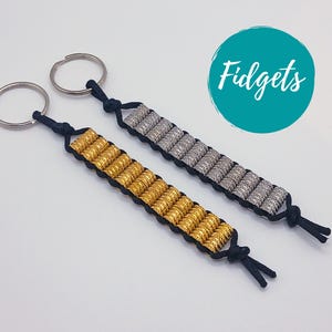 May include: Two beaded fidget keychains. One is gold, the other silver, both with black cord and key rings. The word "Fidgets" is in white script on a teal circle in the background. These keychains are designed to help with anxiety.