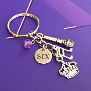 May include: A silver-toned metal keyring featuring a circular ring attached to four charms: a small purple glitter bead, a round disc engraved with the word SIX, a musical note symbol, a crown, and a miniature microphone charm.