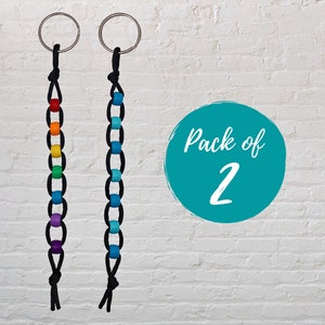 May include: Two black string keychains with rainbow coloured beads. The keychains are hanging against a white brick wall. The text "Pack of 2" is written on a teal circle.