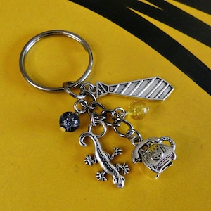 May include: Silver-coloured keyring with a tie charm, a lizard charm, and a rotary phone charm. The keyring also features a dark blue speckled bead and a yellow bead. The background is yellow with black stripes.