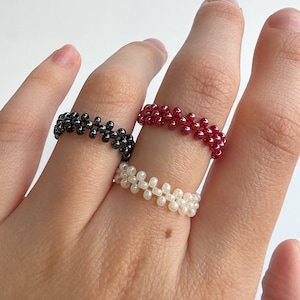 Könnte beinhalten: Drei Perlenringe an einer Hand. Die Ringe bestehen aus kleinen schwarzen, weißen und roten Perlen.