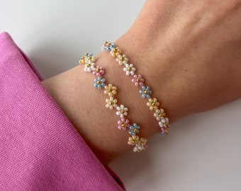 May include: Two beaded bracelets with a daisy flower design. The flowers are white, pink, yellow, and light blue with gold centers. The bracelets are worn on a wrist, with a pink sleeve in the background.