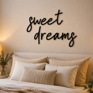 May include: A bedroom scene featuring a bed with multiple pillows and a brown blanket. Above the bed, a black cursive sign reads "sweet dreams". A lamp and greenery are visible on a nightstand. The overall color scheme is neutral.