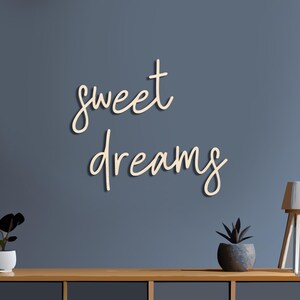 May include: A wooden wall art piece with the words "sweet dreams" in a cursive font. The letters are a light beige color, and the wall behind it is a muted blue. A wooden table holds a small plant and a lamp.
