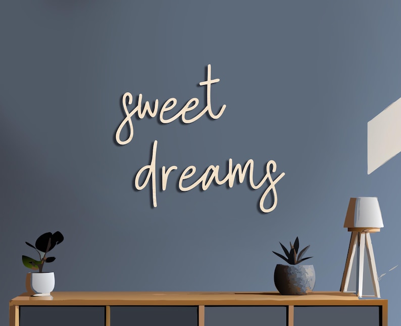 May include: A wooden wall art piece with the words "sweet dreams" in a cursive font. The letters are a light beige color, and the wall behind it is a muted blue. A wooden table holds a small plant and a lamp.
