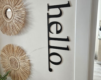 May include: Black metal "hello" sign mounted vertically on a white wall. Two woven wall decorations with a sunburst design are also visible. The sign is in a modern font and adds a decorative touch to the space.