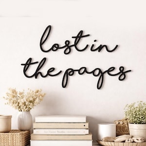 May include: Black wooden wall art with the words "lost in the pages" in a cursive font. The art is mounted on a white wall above a display of books, vases, and decorative items.