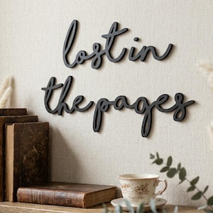 Puede incluir: Arte de pared de madera negra con la frase "lost in the pages" en escritura cursiva. El arte está montado en una pared de color neutro sobre una estantería con libros antiguos y una taza de té.
