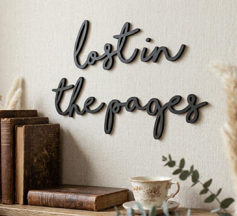 May include: Black wooden wall art spelling "lost in the pages" in cursive script. The art is mounted on a neutral-colored wall above a shelf with antique books and a teacup.