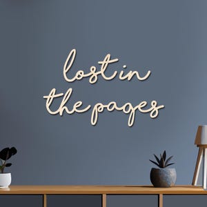 May include: Wooden wall art with the phrase "lost in the pages" in a cursive font. The text is light beige and mounted on a blue wall. A wooden table with plants and a lamp is below the text.