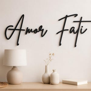 May include: Black cursive wall art spelling "Amor Fati" on a white wall. The words are above a beige lamp, two small vases, and a closed book on a wooden surface. The lamp has a round base and a cylindrical lampshade.
