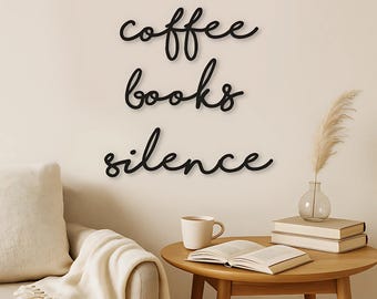 Puede incluir: Letras de pared en cursiva negra que forman las palabras coffee, books y silence en tres líneas separadas. Las letras tienen un acabado oscuro y liso, y están dispuestas verticalmente sobre una superficie de pared de color claro.
