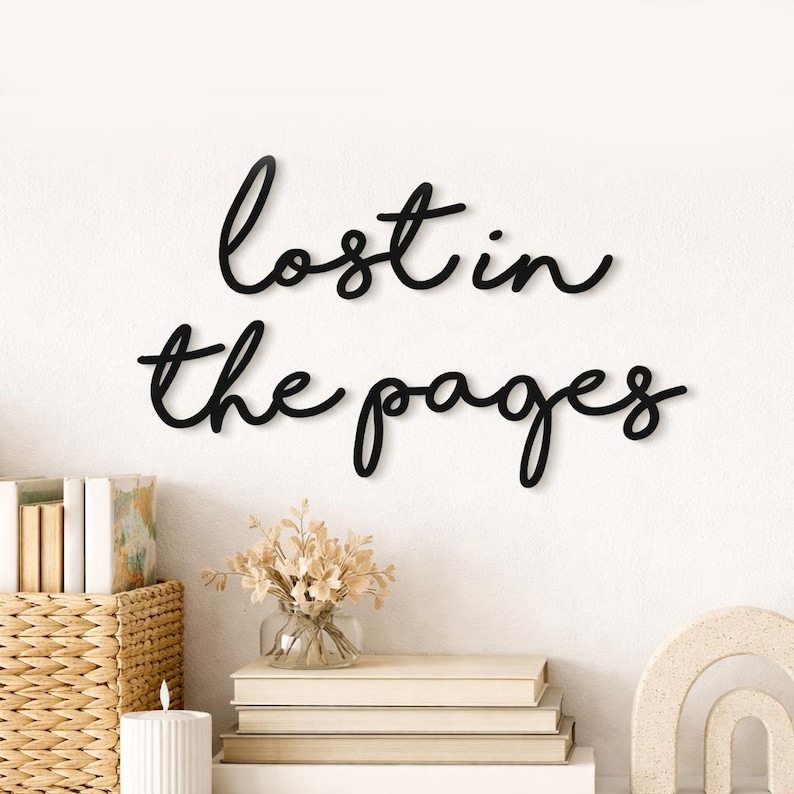 May include: Black cursive wall art that reads "lost in the pages" is mounted on a white wall. The decor includes a woven basket, books, a candle, and a vase of dried flowers. The overall theme is home decor.