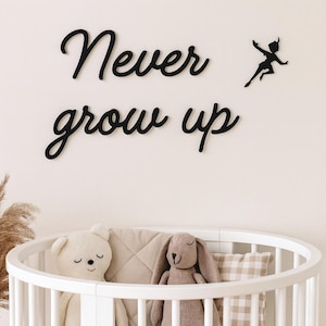 Könnte beinhalten: Schwarze Wanddekoration aus Holz mit dem Schriftzug "Never grow up" in Schreibschrift und einer Silhouette von Peter Pan. Im Vordergrund befindet sich ein weißes Babybett mit Stofftieren und Kissen. Die Stofftiere umfassen einen Bären und ein Kaninchen.