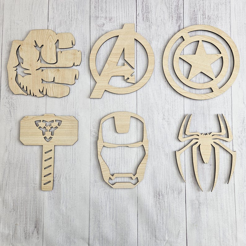 K&ouml;nnte beinhalten: Ein Set aus sechs hellen Holzausschnitten mit verschiedenen Superhelden-Motiven, darunter eine geballte Faust, der Buchstabe A, ein Stern in einem Kreis, ein Hammer, eine Helmmaske und eine Spinnensilhouette, angeordnet auf einer wei&szlig;en Holzoberfl&auml;che.