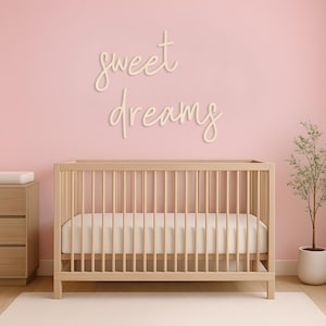 May include: A nursery scene featuring a light wood crib with a white mattress and a dresser. The pink wall has the words "sweet dreams" in cursive. A small potted plant sits to the right, and a white rug is on the floor.