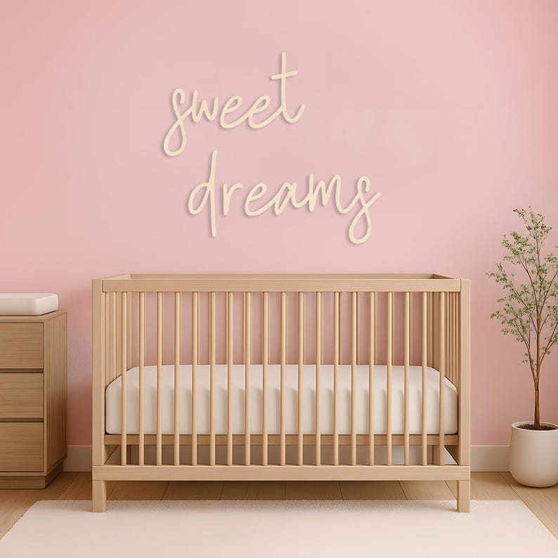 May include: A nursery scene featuring a light wood crib with a white mattress and a dresser. The pink wall has the words "sweet dreams" in cursive. A small potted plant sits to the right, and a white rug is on the floor.