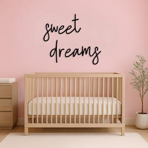 May include: A nursery scene featuring a light wood crib with a white mattress against a pink wall. Above the crib, the words "sweet dreams" are written in a flowing black script. A small dresser and a potted plant complete the room's decor.
