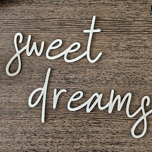 May include: Silver metal wall art spelling out "sweet dreams" in a cursive font. The words are placed on a dark brown wooden surface. The letters are elegant and modern, suitable for bedroom decor.