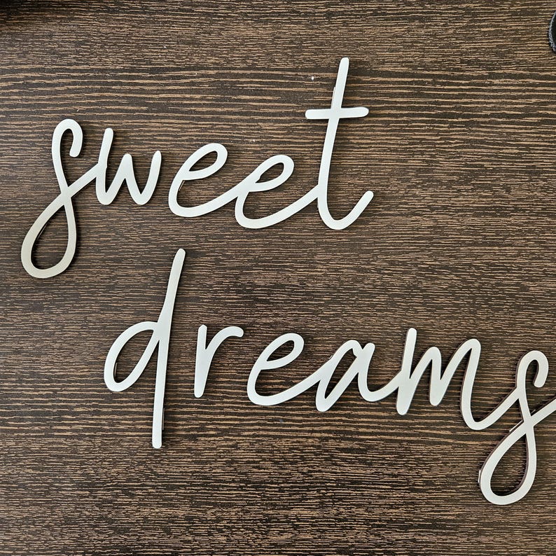 May include: Silver metal wall art spelling out "sweet dreams" in a cursive font. The words are placed on a dark brown wooden surface. The letters are elegant and modern, suitable for bedroom decor.