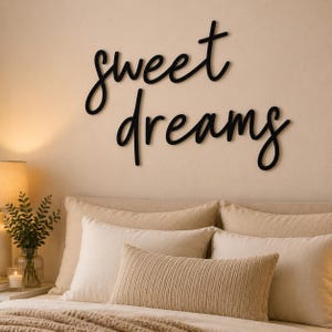 May include: Black cursive "sweet dreams" wall art above a bed with beige pillows and a textured throw. A lamp and greenery in a vase are on the bedside table. The wall is a light beige colour.