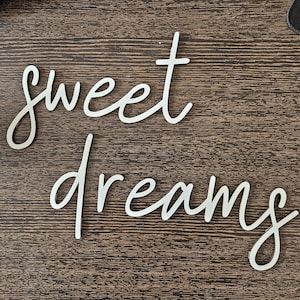 May include: White cursive wooden sign that reads "sweet dreams" on a dark brown wooden surface. The words are in a flowing, elegant script, perfect for bedroom decor.