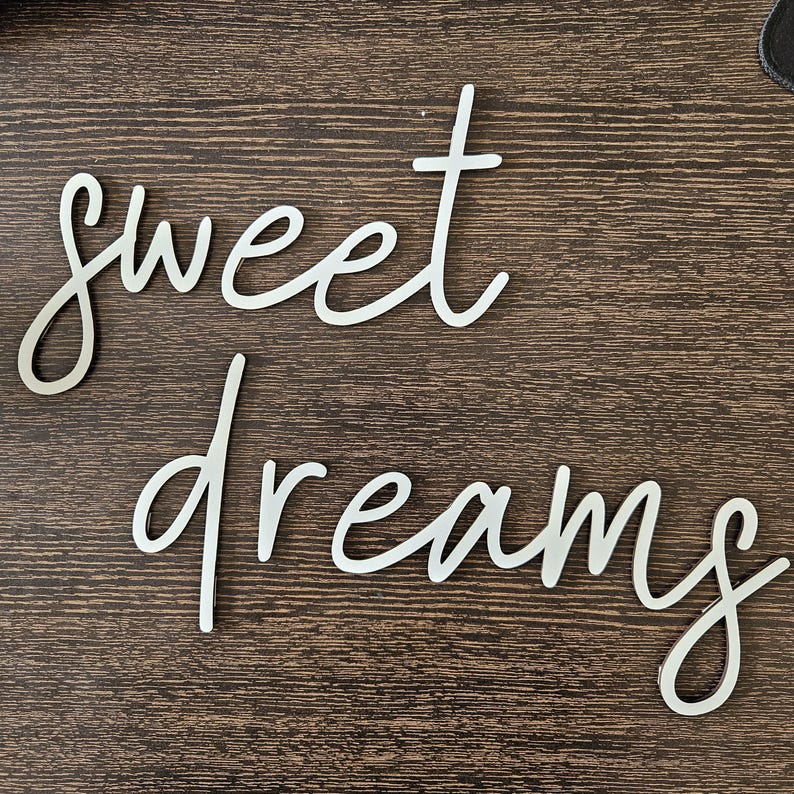 May include: White cursive wooden sign that reads "sweet dreams" on a dark brown wooden surface. The words are in a flowing, elegant script, perfect for bedroom decor.