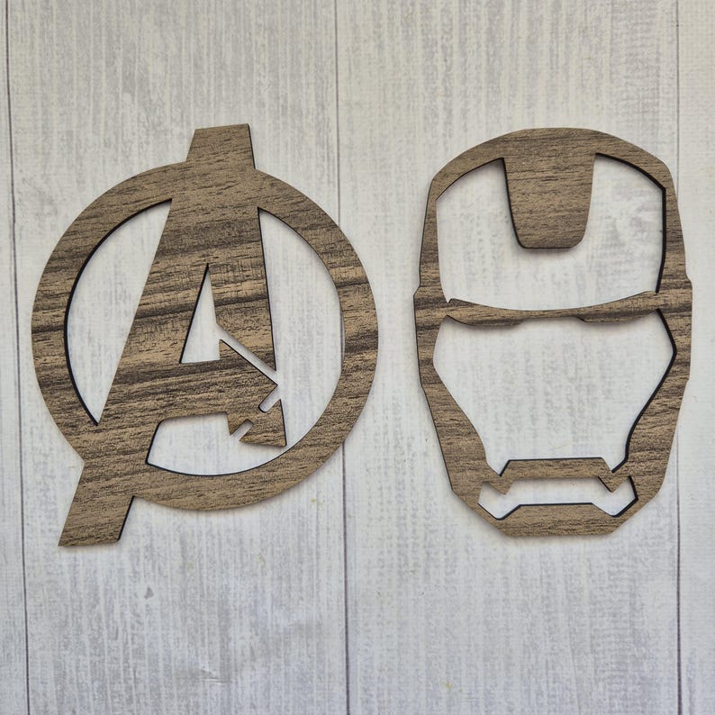 K&ouml;nnte beinhalten: Zwei flache, lasergeschnittene Wanddekorationen aus Holz mit brauner Holzmaserung. Ein Teil ist das Avengers-Logo, ein gro&szlig;er Buchstabe A in einem Kreis. Das zweite Teil ist die stilisierte Maske der Figur Iron Man mit Aussparungen.