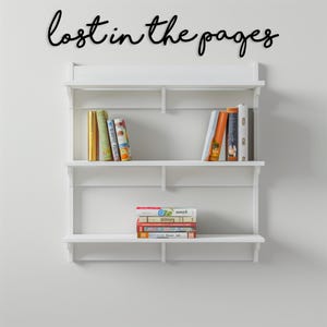May include: A white wall-mounted bookshelf with three shelves, holding a collection of books. The phrase "lost in the pages" is written in black script above the shelves. The books have colorful spines.