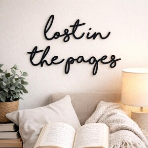 May include: Black cursive wall art that reads "lost in the pages" is mounted on a white wall. A cozy reading nook is set up below with a book, pillows, a blanket, and a lamp. A potted plant in a woven basket sits to the left.