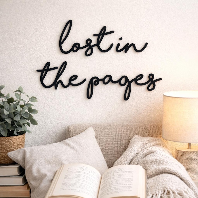 May include: Black cursive wall art that reads "lost in the pages" is mounted on a white wall. A cozy reading nook is set up below with a book, pillows, a blanket, and a lamp. A potted plant in a woven basket sits to the left.