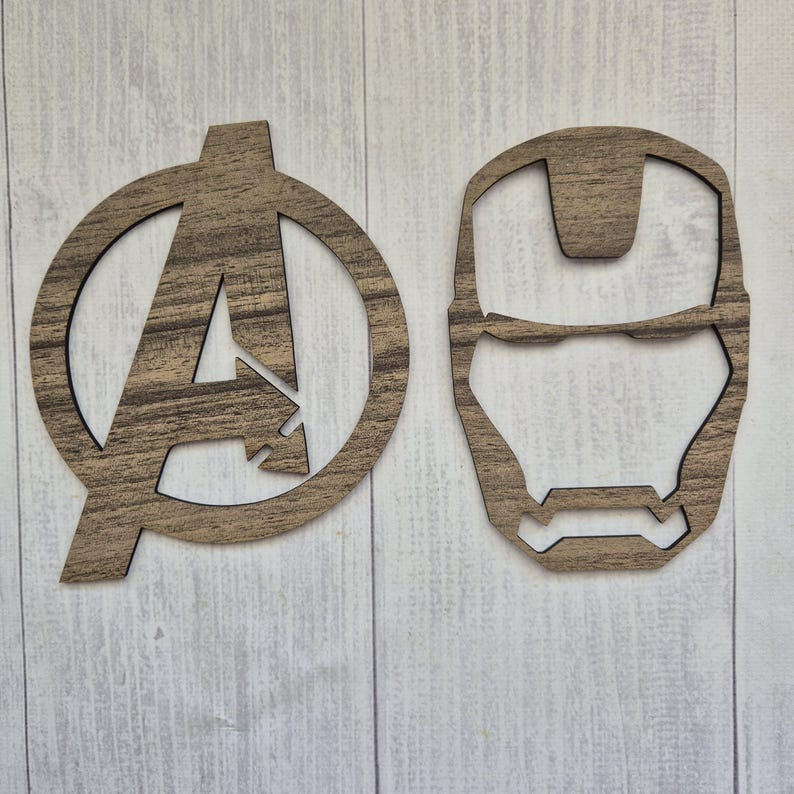 K&ouml;nnte beinhalten: Zwei flache, lasergeschnittene Wanddekorationen aus Holz mit einer dunkelbraunen Holzmaserung. Ein Teil ist das kreisf&ouml;rmige Avengers-Logo mit einem zentralen A, das andere ist der stilisierte Umriss der Iron Man-Maske.