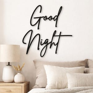 May include: Black cursive "Good Night" wall art. The words are displayed on a white wall above a bed with neutral-toned bedding. A white lamp and a small vase with dried flowers sit on a wooden nightstand.