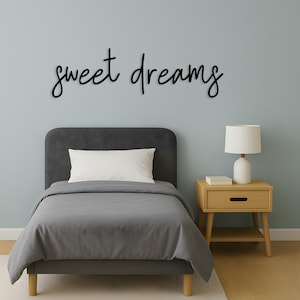 May include: A bedroom scene featuring a gray bed with a white pillow and gray bedding. Above the bed, the words "sweet dreams" are written in black script. A wooden nightstand with a lamp sits beside the bed.