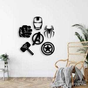 K&ouml;nnte beinhalten: Eine Ansammlung von sechs schwarzen Wanddekorationen, bestehend aus einer Superheldenmaske, einer geballten Faust, einem Spinnenemblem, dem Avengers-Logo, einem Hammer und einem runden Schild mit einem Stern in der Mitte, angeordnet an einer wei&szlig;en Wand.