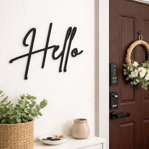 Pode incluir: Decoração de parede preta com a palavra "Hello" em escrita cursiva. A palavra está montada em uma parede branca. Uma cesta de vime com vegetação, um vaso pequeno e um prato decorativo estão em uma prateleira branca abaixo.
