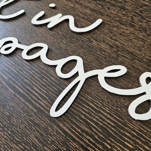 May include: White cursive lettering spelling "in pages" on a dark brown wooden surface. The letters are cut out and appear to be made of a smooth material. The image is a close-up shot.