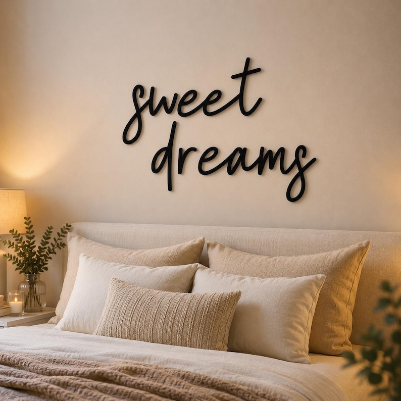 May include: A bedroom scene featuring a bed with multiple pillows and a brown blanket. Above the bed, a black cursive sign reads "sweet dreams". A lamp and greenery are visible on a nightstand. The overall color scheme is neutral.