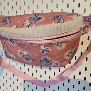 Könnte beinhalten: Rosa Bauchtasche mit einem sich wiederholenden Muster einer Zeichentrickfigur in Blau und Weiß. Die Tasche hat eine weiße Cord-Borte und einen rosa Riemen. Eine schwarze Handschlaufe ist an der Seite befestigt.