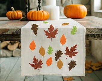 May include: A white table runner with a fall leaf design. The runner features orange, brown, and green leaves in various shapes and sizes. The runner is laid out on a wooden table with pumpkins and candles.