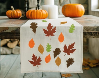 May include: A white table runner with a fall leaf design. The runner features orange, brown, and green leaves in various shapes and sizes. The runner is laid out on a wooden table with pumpkins and candles.