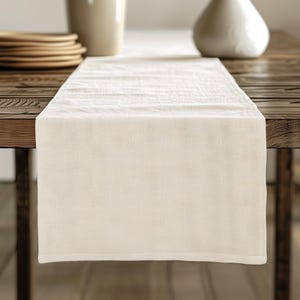 May include: A white linen table runner with a subtle texture. The runner is laid out on a wooden table.