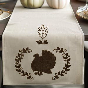 May include: A beige table runner with a brown silhouette of a turkey surrounded by a wreath of leaves. Above the turkey is a brown silhouette of a leaf.