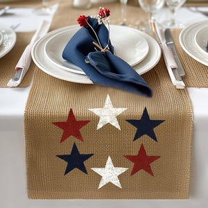 May include: A burlap table runner with red, white, and blue star designs. The runner is placed on a white tablecloth and is part of a table setting with white plates, blue napkins, and silverware. The table setting has a patriotic theme.