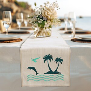 May include: A white table runner with a tropical design featuring palm trees, a dolphin, and a seagull. The design is in shades of teal and green on a beige background.