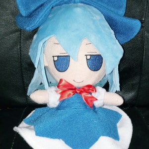 Puede incluir: Un juguete de peluche azul con un lazo azul, un lazo rojo y un vestido blanco. El juguete tiene una cara sonriente con ojos azules.